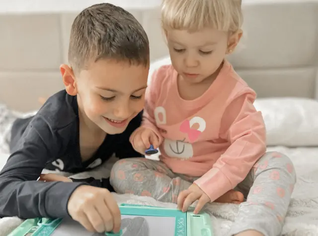 Dwoje małych dzieci siedzi na łóżku, bawiąc się radośnie tabletem do rysowania i ze sobą nawzajem, otoczone miękką pościelą.