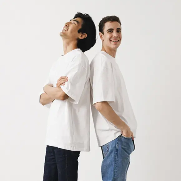 Dwóch młodych mężczyzn w białych koszulkach stoi tyłem do siebie na białym tle i uśmiecha się swobodnie.