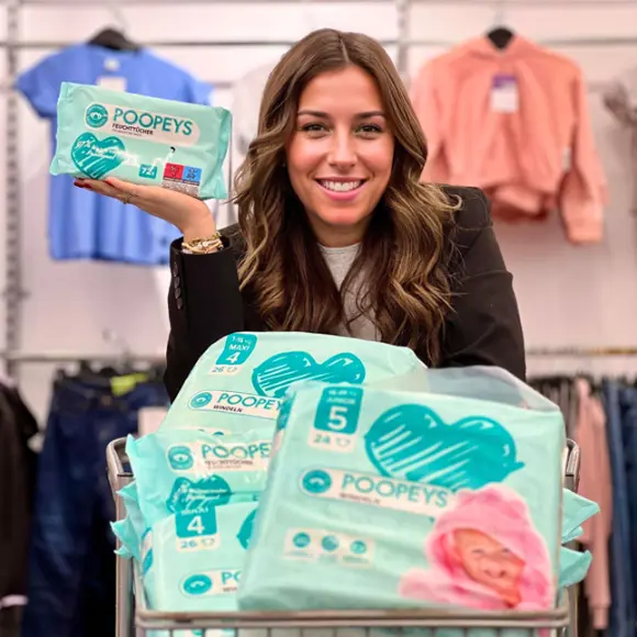 Osoba trzymająca paczkę chusteczek nawilżanych Poopeys, otoczona ułożonymi w stos paczkami