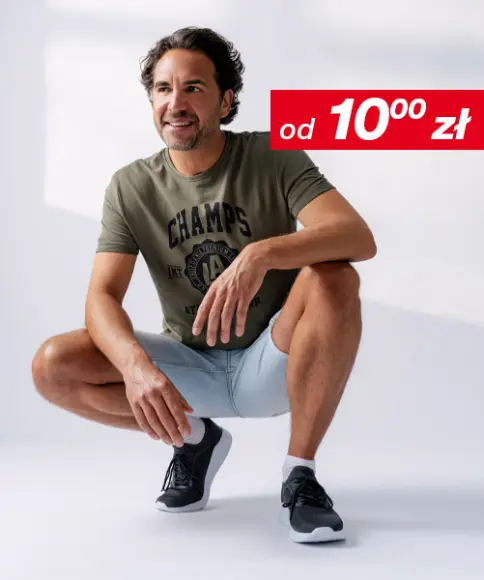 Mężczyzna w swobodnej pozie, ubrany w zielony T-shirt i jasnoniebieskie szorty, w czarnych butach sportowych, na tle miękkiego tła.