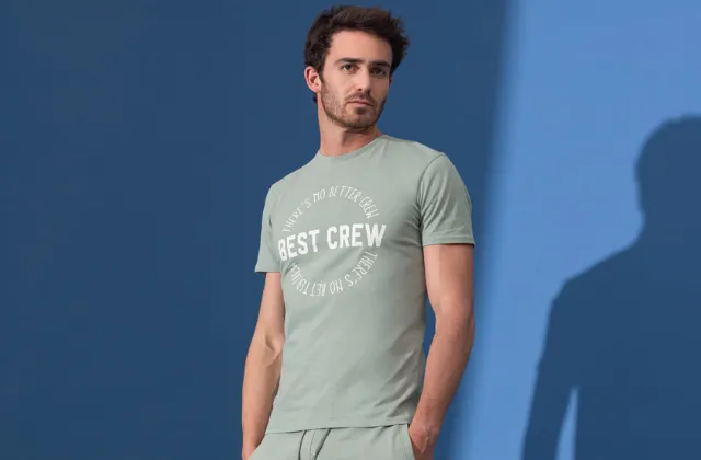 Osoba w jasnozielonym T-shircie z białym napisem „BEST CREW” pozuje na tle niebieskiego tła. Swobodny i stylowy wygląd.