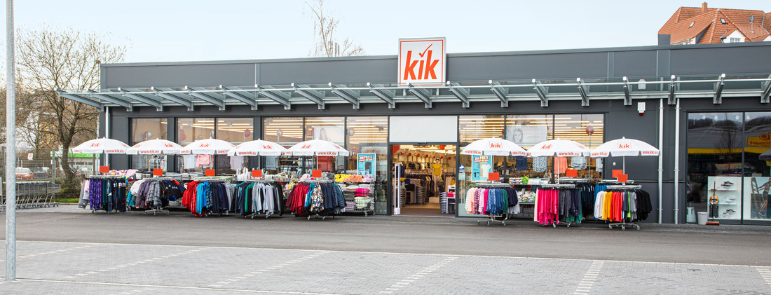 Widok zewnętrzny sklepu KiK z logo KiK na drzwiach i stojakami z ubraniami przed wejściem