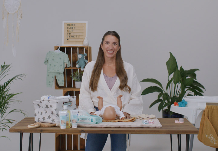 Kobieta stoi przy drewnianym stole z produktami do pielęgnacji niemowląt, ubraniami i zabawkami, otoczona zielenią w jasnym otoczeniu. Kobieta stoi przy drewnianym stole z produktami do pielęgnacji niemowląt, ubraniami i zabawkami, otoczona zielenią w jasnym otoczeniu.
