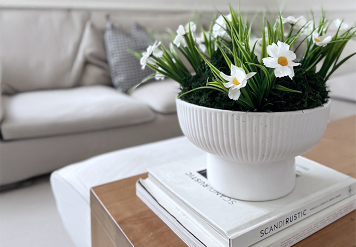 Biała miska z sztucznymi stokrotkami i zielenią stoi na ułożonych w stos książkach na drewnianym stoliku kawowym, w tle jasna sofa.