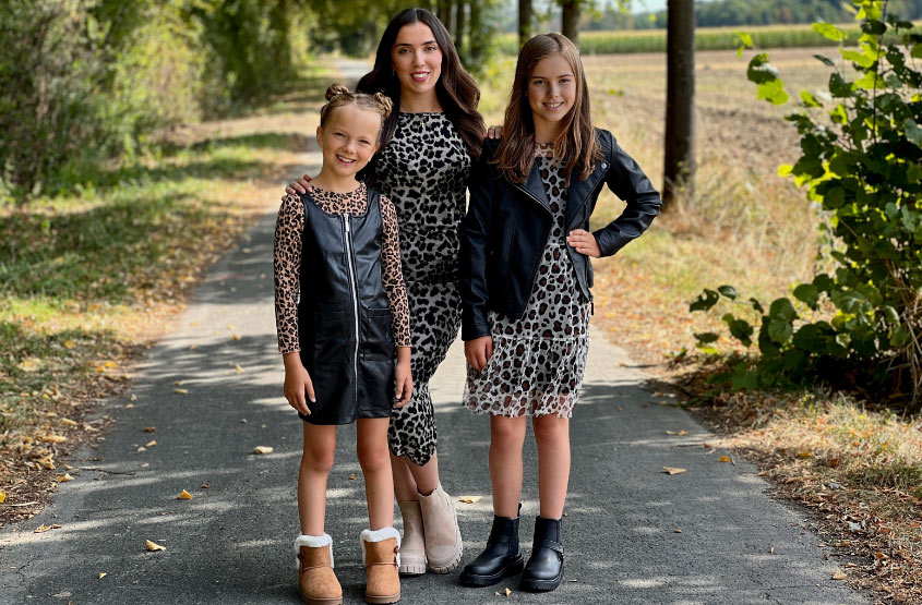 Drei Kinder posieren auf einem mit Blättern übersäten Weg, gekleidet in stilvolle Outfits mit Leopardenmuster, umgeben von Bäumen und einem klaren Himmel über ihnen.