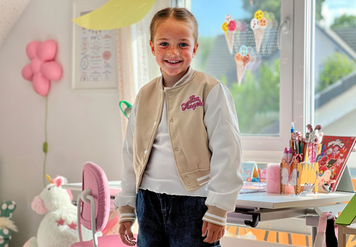 Dziecko w stylowej beżowo-białej kurtce typu varsity stoi w kolorowym, radosnym pokoju pełnym przyborów plastycznych i ozdób.