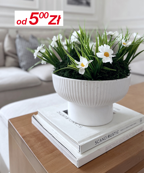 Biała miska z sztucznymi stokrotkami i zielenią stoi na ułożonych w stos książkach na drewnianym stoliku kawowym, w tle jasna sofa.